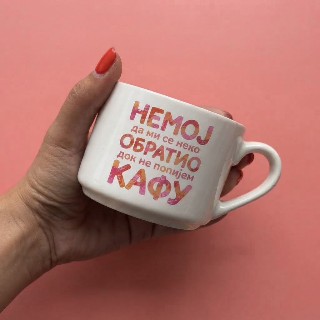 Bela šoljica u ruci sa šarenim natpisom Nemoj da mi se neko obratio dok ne popijem kafu